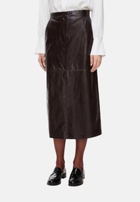Gonna midi in pelle marrone con una texture liscia, caratterizzata da una chiusura frontale con bottoni e cuciture verticali. Modello che indossa mocassini neri.