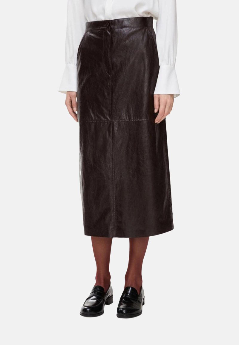 Gonna midi in pelle marrone con una texture liscia, caratterizzata da una chiusura frontale con bottoni e cuciture verticali. Modello che indossa mocassini neri.
