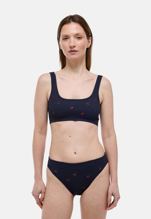 Femme portant un bikini côtelé bleu marine avec de petits motifs de cerises rouges, debout devant un fond clair uni.