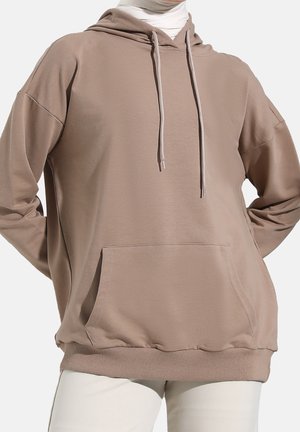 Beiger Oversize-Hoodie mit Fronttasche und Kordelzug, getragen über einem weißen Rollkragenpullover und kombiniert mit weißen Hosen.