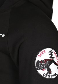 Mustapaitinen huppari, jossa on pyöreä brodeerattu merkki mustasta pantterista, punaisista yksityiskohdista ja tekstistä "HAE ISKE TUHOA". Silkkinen pinta.