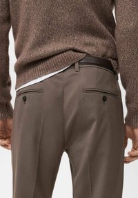 Suéter de lana marrón sobre una camisa gris claro, combinado con pantalones marrones de corte tailored. Los pantalones cuentan con dos bolsillos traseros con cierres de botón.