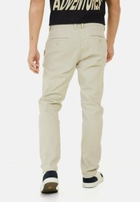Pantaloni beige chiari a taglio dritto, con due tasche posteriori con chiusura a bottone, indossati con sneakers blu navy e una camicia nera.