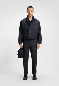 Mørk denimjakke med cropped snit, to brystlommer, parret med matchende mørke denimbukser og sorte støvler, mens der holdes en lille sort taske.
