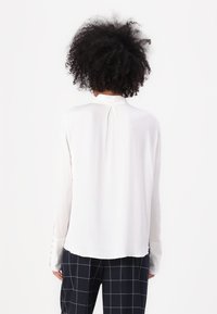 Blusa blanca de manga larga con cuello alto, textura suave y puños abotonados. Presenta un pliegue en la parte posterior para un detalle de diseño adicional.