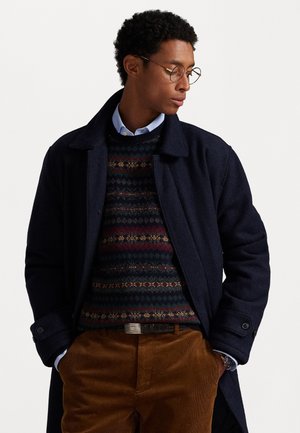 Manteau en laine bleu marine, pull à motifs avec des accents verts, rouges et dorés, pantalon en velours côtelé marron et chemise habillée grise avec boutons.