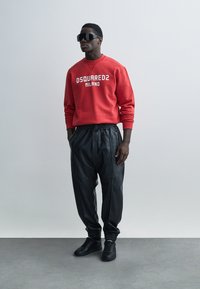 Κόκκινο φούτερ με το λογότυπο "DSQUARED2 MILANO", σε συνδυασμό με μαύρο δερμάτινο παντελόνι jogger με ελαστικές μανσέτες και μαύρα αθλητικά παπούτσια.