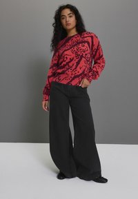 Rot und schwarz gemusteter Strickpullover mit lockerer Passform, kombiniert mit weiten schwarzen Hosen und schwarzen flachen Schuhen vor einem schlichten Hintergrund.