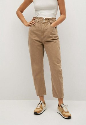 Personne portant un pantalon beige taille haute, un top blanc sans manches et des baskets beige et blanches, debout les mains dans les poches.