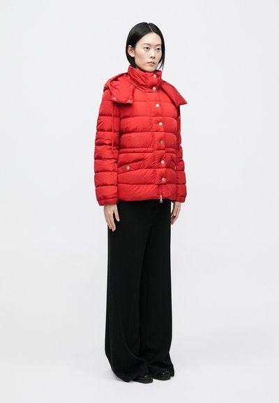 Frau steht in einer leuchtend roten Steppjacke mit Druckknöpfen und weiten schwarzen Hosen vor einem schlichten hellen Hintergrund.