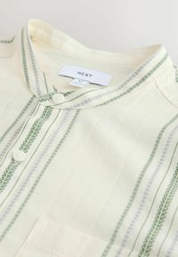 Koszula z krótkim rękawem w jasnopółżółtej tkaninie, z zielonymi i szarymi pionowymi paskami, zapinana na guziki, z kołnierzykiem i kieszeniami.