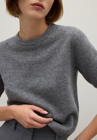 Suéter gris de punto con cuello redondo, mangas cortas y una textura suave, con una mano descansando en el dobladillo, frente a un fondo liso.