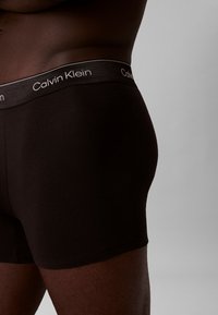 Schwarze Baumwoll-Boxershorts mit einem grauen Calvin Klein-Logobund, die eine glatte Textur und einen figurbetonten Schnitt aufweisen, seitlich gezeigt.
