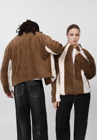 Brun og cremefarvet fleecejakke med lynlås foran, høj krave og kontrasterende paneler. Elastikker ved kant og manchetter, kombineret med sorte jeans.