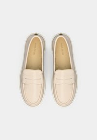 Ζευγάρι μπεζ δερμάτινων μοκασίνων με λουρί για κέρμα και άσπρες σόλες, απεικονισμένα από πάνω σε λευκό φόντο.