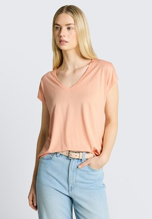 Femme blonde portant un haut pêche à manches courtes col en V, rentré dans un jean bleu clair avec une ceinture beige, posant avec une main dans la poche.