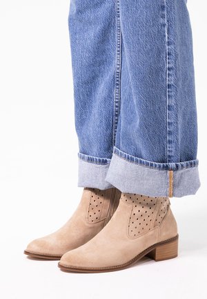 Beige suede enkelboots met een puntige neus en een geperforeerd ontwerp. Lage gestapelde hak, gecombineerd met blauwe jeans met opgerolde zoom.