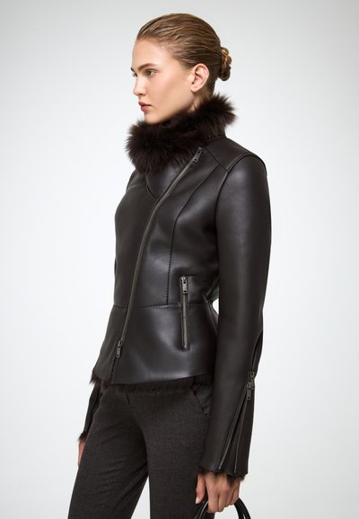 Donna che indossa una giacca di pelle marrone scuro con colletto e polsini in pelliccia, tasche con zipper e pantaloni marroni, mentre tiene una borsa nera.