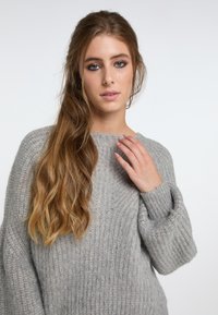 Pulover gri tricotat, cu o croială lejeră, textură canelată și mâneci late cu manșete. Modelul poartă părul lung și ondulat, cu mâna sprijinită pe umăr.