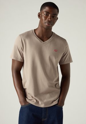 Homme portant un t-shirt beige à col en V Levi’s et un jean bleu foncé, les mains dans les poches, devant un fond clair uni.