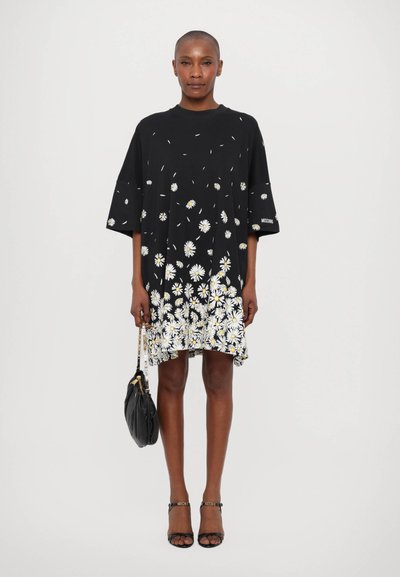 Sort kjole med hvidt og gult tusindfrydsmønster, korte ærmer, afslappet pasform og kant med blomstergrafik; accessoriseret med en sort taske.