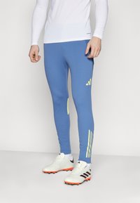 Blue athletic leggings featuring a fitted design, soft fabric, yellow accents, and three vertical stripes on the lower leg. Pair of white sneakers.