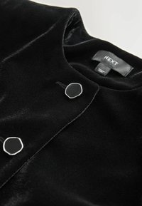 Black velvet jacket featuring a round neckline, silver hardware, and a smooth texture. Label stitched on the inside, showing brand name.