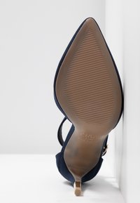 Chaussure à talon haut bleu marine unique avec bout pointu et semelle texturée, vue de dessous sur fond blanc.