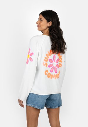 Woman with long dark hair wearing white sweatshirt with pink flower and orange text "SUNDAYS COMING" on back and denim shorts.