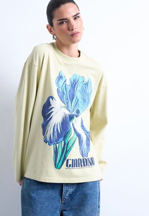 Personne portant une chemise beige à manches longues avec un grand motif fleur bleu et blanc et le texte "GIARDINO", associée à un jean bleu.