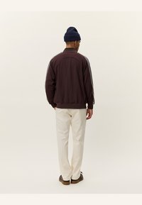 Sacou sport maro cu dungi laterale albe, guler și manșete tivite, asortat cu pantaloni crem și o căciulă albastră.