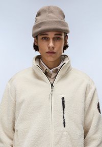 Jachetă din blană artificială în nuanță crem, cu guler înalt, fermoar frontal, detalii de buzunare negre și o textură moale. Căciulă maro purtată pe cap.