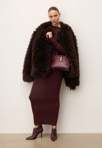 Un vestido ajustado burdeos combinado con un abrigo de piel shaggy marrón. La modelo sostiene un bolso burdeos con herrajes dorados. Botas de tacón alto y puntera.