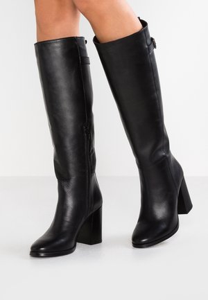 Bottes en cuir noires montantes jusqu'au genou avec talons carrés portées sur jambes nues, avec fermetures éclair latérales et détails de boucles près du haut.