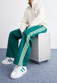 Gröna träningsbyxor med gula sidostickningar, tillsammans med en krämfärgad zip-up sweatshirt och vita sneakers med gröna accenter.