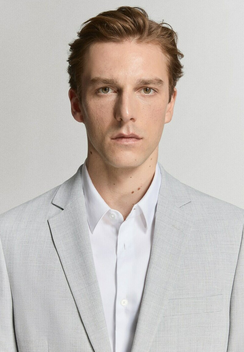 Jonge man met lichtbruin haar, gekleed in een lichtgrijze blazer en een wit overhemd, neutrale uitdrukking, grijze achtergrond.
