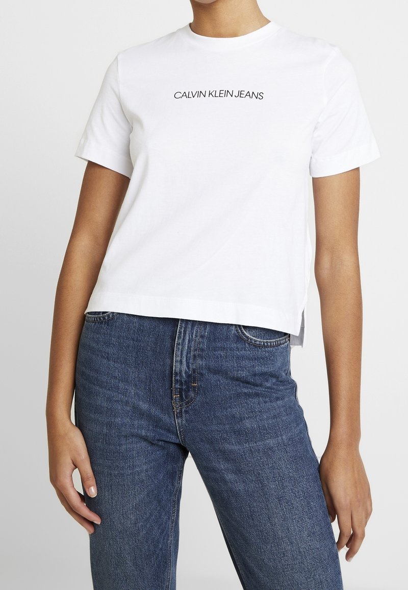 Biały bawełniany t-shirt z okrągłym dekoltem i krótkimi rękawami, z czarnym nadrukiem "CALVIN KLEIN JEANS" na wysokości klatki piersiowej.