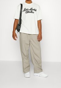 Uomo che indossa una maglietta bianca "Low Lights Studios", pantaloni beige larghi, sneakers bianche e porta una borsa a tracolla nera su uno sfondo bianco.