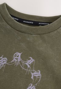 Olivgrön bomulls-t-shirt med rund halsringning, med en ljuslila abstrakt grafisk design på framsidan och en texturerad yta.