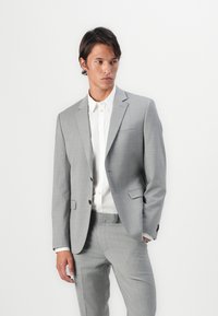 Light grey tailored suit with a smooth texture, single-breasted blazer, notch lapels, and matching trousers. White button-up shirt underneath.