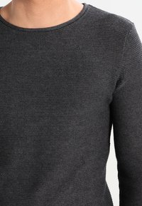 Pull en maille gris foncé avec un col rond et des manches longues, présentant un tissu texturé et un design ajusté.