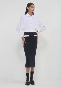 Donna in camicia bianca a maniche lunghe con bottoni, infilata in una gonna midi nera a vita alta con alette delle tasche bianche, calze nere e mocassini.