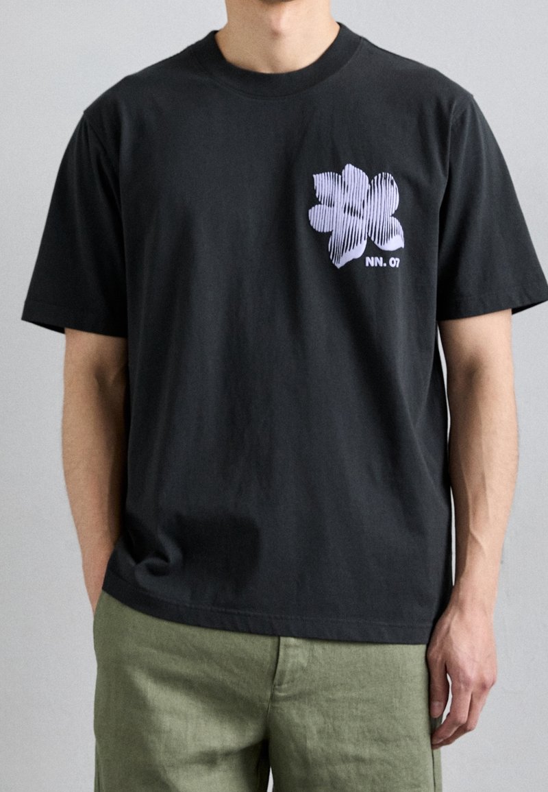 Homme portant un T-shirt noir à manches courtes avec une fleur rayée blanche et le texte "NN. 07", associé à un pantalon vert clair, se tenant devant un mur gris.