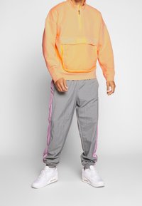 Pullover arancione neon con mezza zip e tasca frontale; pantaloni della tuta grigi con strisce laterali rosa; sneakers bianche completano l'outfit.