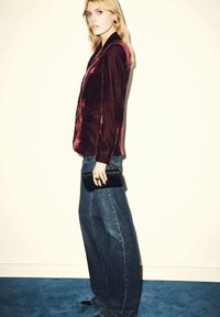 Blazer de terciopelo burdeos con solapas, combinado con jeans de mezclilla de tiro alto y pierna ancha. Un clutch negro y unos zapatos de punta negra completan el look.