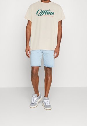 Camiseta beige con gráfico "Offline" en verde y rojo, pantalones cortos de mezclilla clara y zapatillas grises con cordones blancos y detalle de logo.