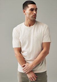 T-shirt beige a maniche corte con colletto rotondo, caratterizzato da un logo discreto sul petto. Tessuto morbido e leggero con una leggera texture.
