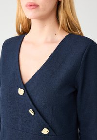 Robe texturée bleu marine avec un col en V, trois boutons dorés sur le devant croisé et des manches longues.