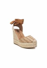 Sandalia de cuña con parte superior de lino beige trenzado, cintas de tela marrón en el tobillo y una suela de estilo esparto texturizada. Punta redondeada con tapa de dedo en contraste.