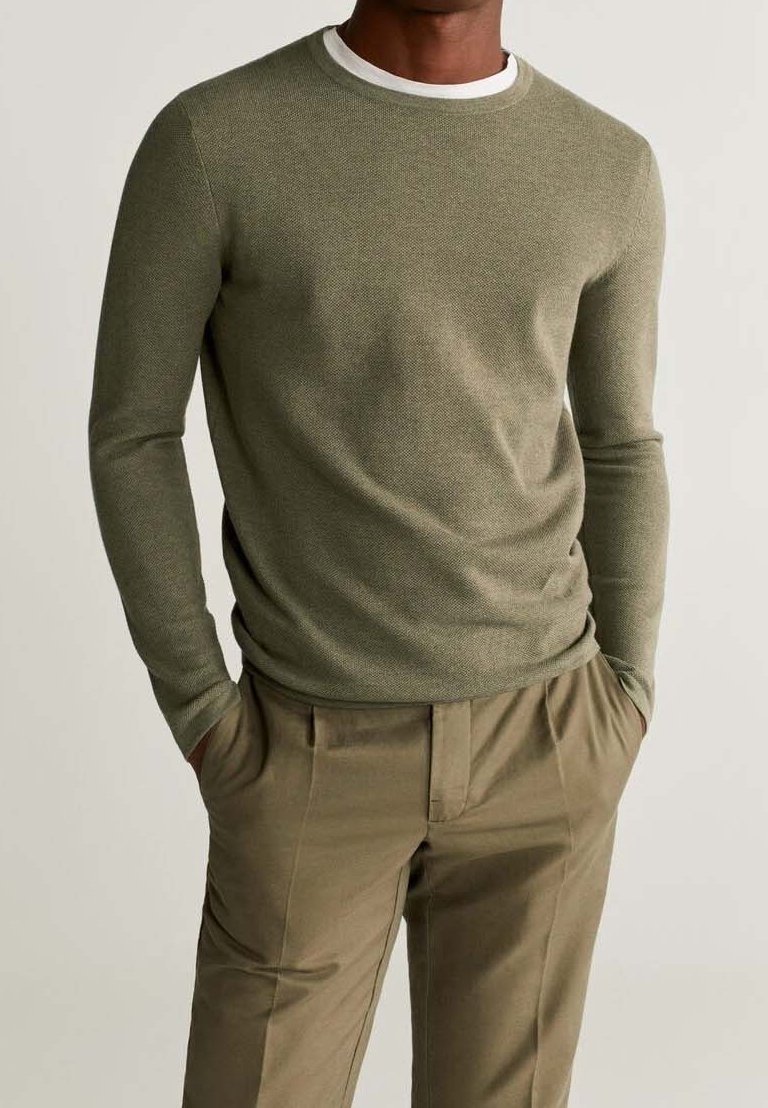 Homme portant un pull texturé à manches longues vert olive sur un sous-vêtement blanc et un pantalon beige, mains dans les poches, fond neutre.
